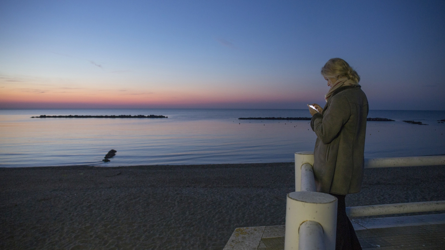 A person wearing a winter coat standing on a seaside promenade at dusk, illuminated by the light of their phone while facing a calm ocean and a sky that transitions from deep blue to soft pink near the horizon. A person wearing a winter coat standing on a seaside promenade at dusk, illuminated by the light of their phone while facing a calm ocean and a sky that transitions from deep blue to soft pink near the horizon.