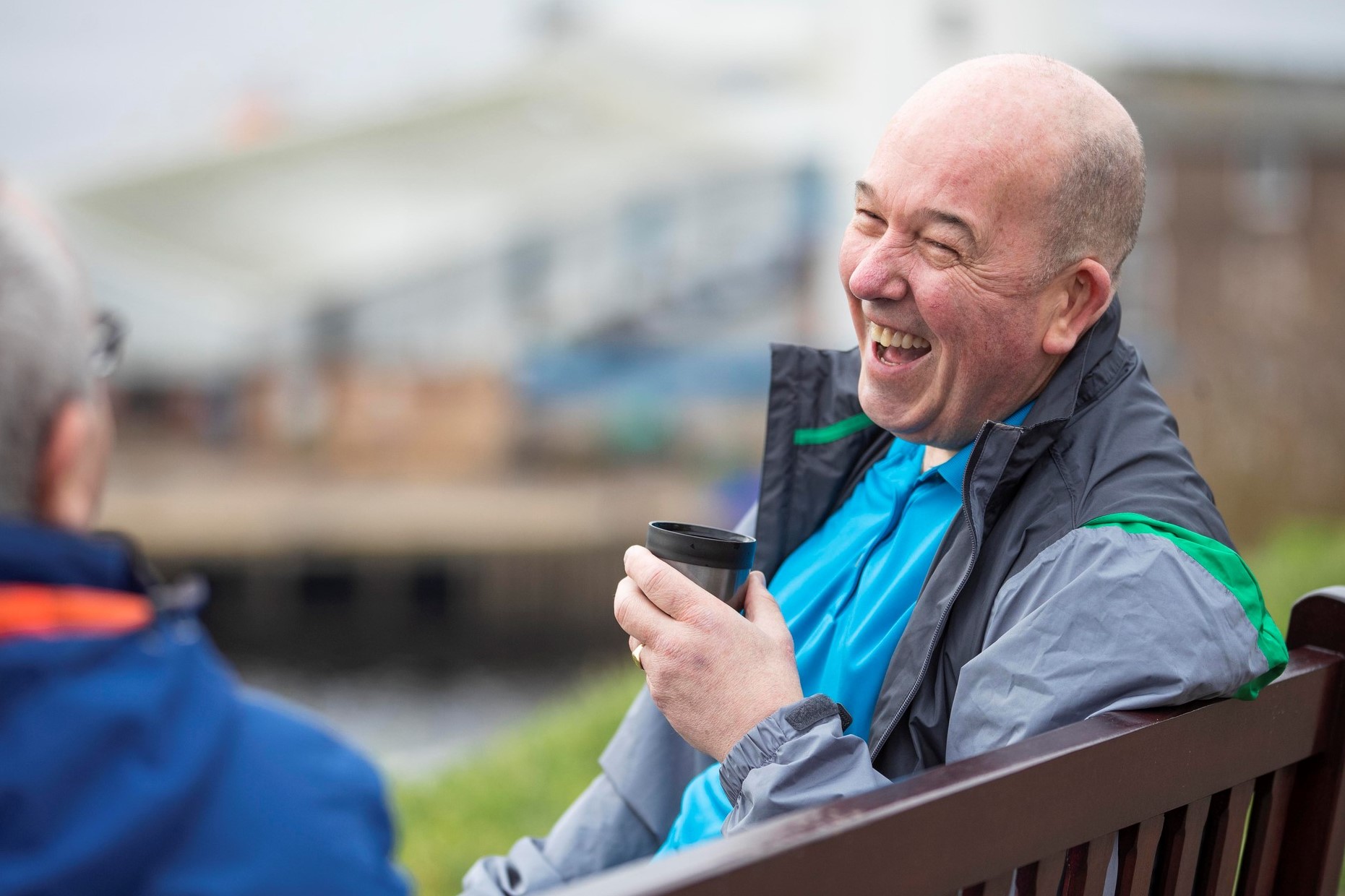 Two people sit on a wooden bench in an outdoor setting. One wears a blue shirt and grey jacket with green accents, holding a black coffee cup.