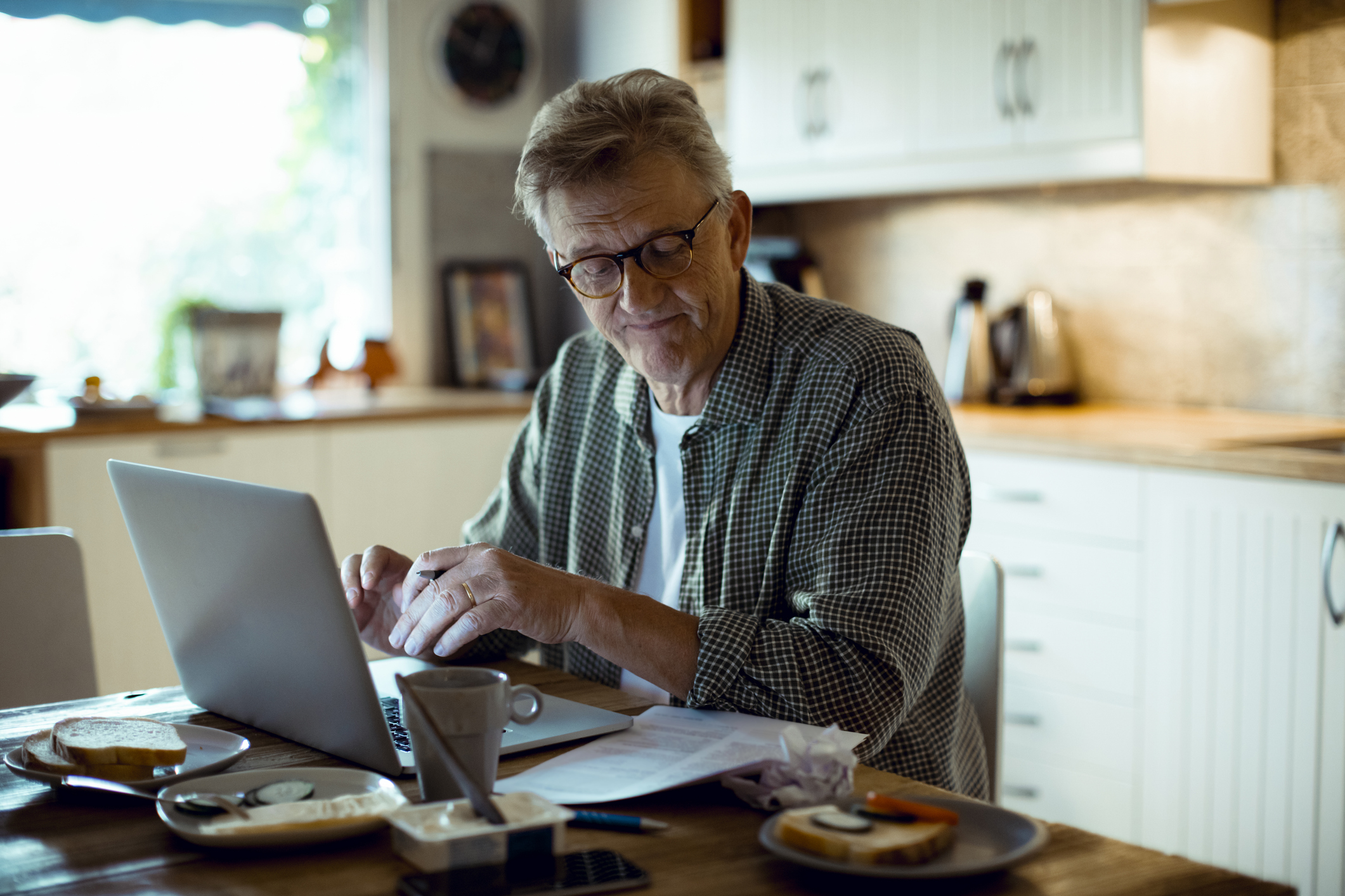 Person sitting at a kitchen table using a laptop, with papers and a coffee cup spread out on the table. The background shows a bright, homey kitchen with white cabinets and a kettle on the counter.