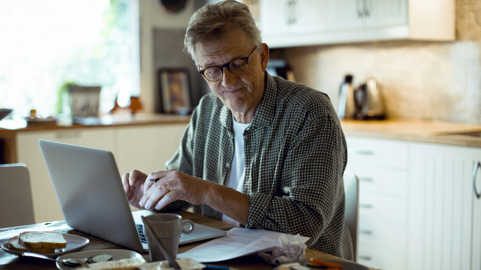 Person sitting at a kitchen table using a laptop, with papers and a coffee cup spread out on the table. The background shows a bright, homey kitchen with white cabinets and a kettle on the counter. Person sitting at a kitchen table using a laptop, with papers and a coffee cup spread out on the table. The background shows a bright, homey kitchen with white cabinets and a kettle on the counter.