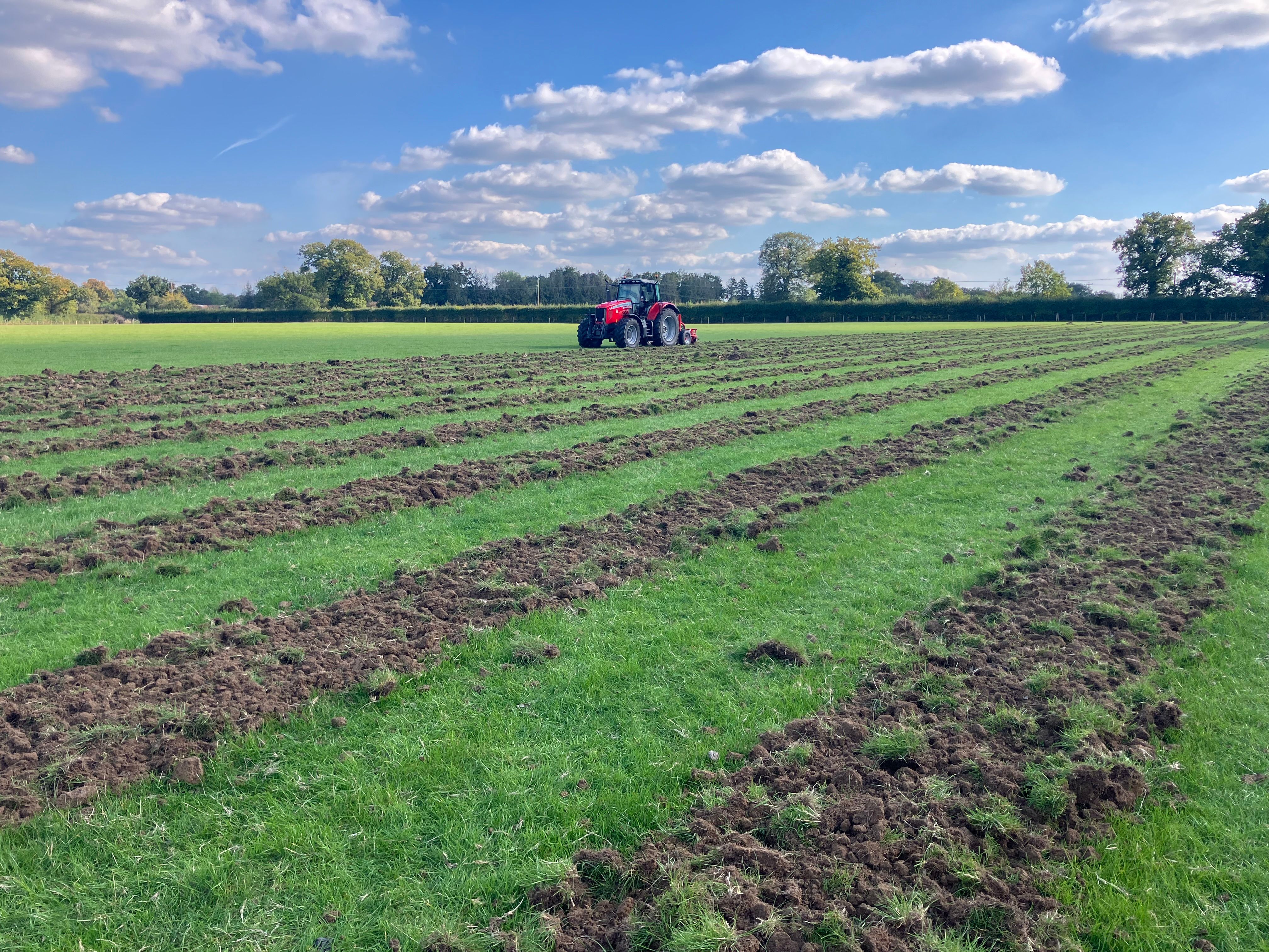 A red tractor working on a large grassy field under a blue sky with scattered white clouds. The field has multiple parallel strips of soil turned over, indicating recent ploughing. Trees and hedgerows line the horizon in the background.
