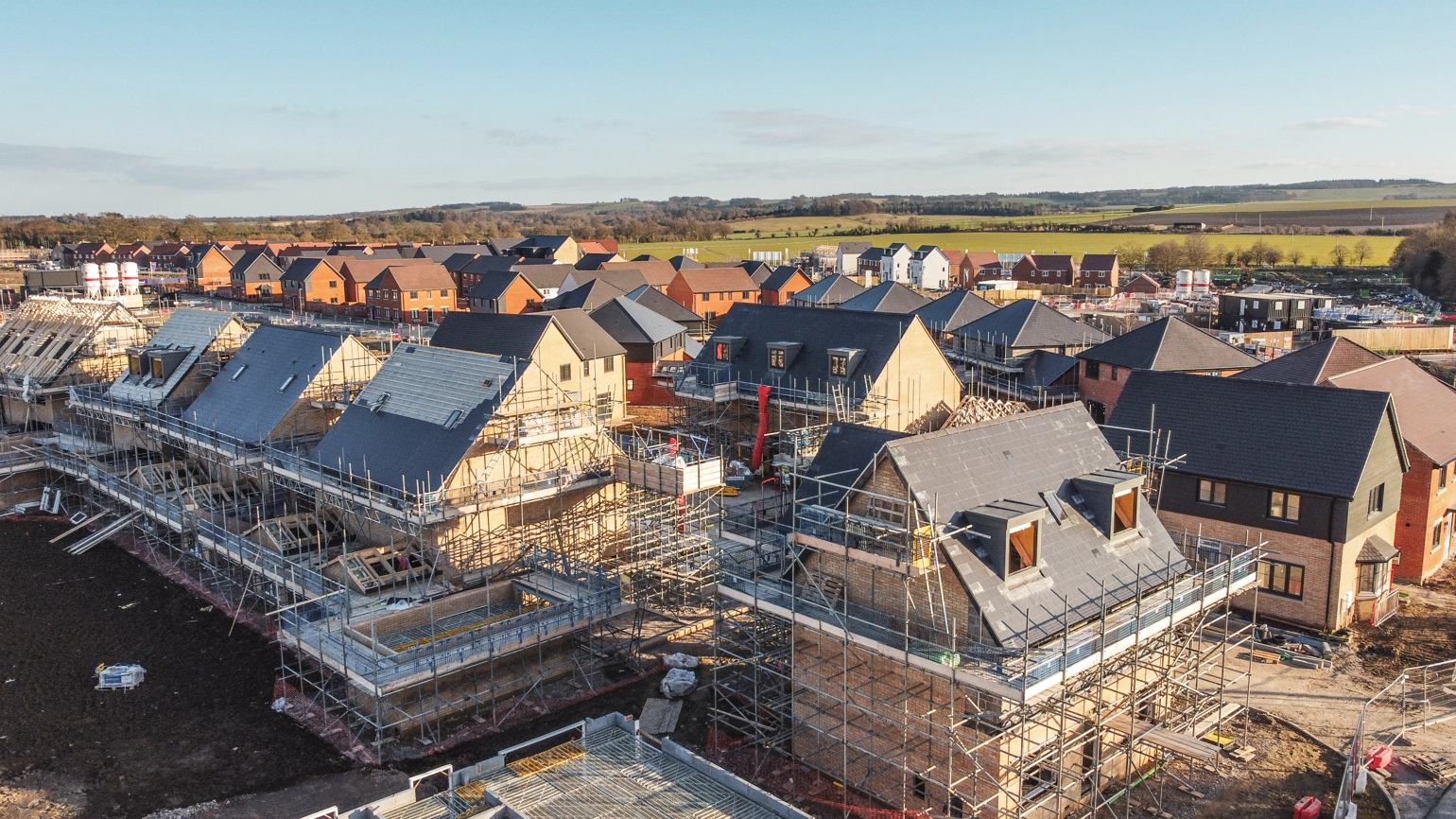 Aerial view of a housing development under construction, with multiple homes in various stages of building and scaffolding visible. Aerial view of a housing development under construction, with multiple homes in various stages of building and scaffolding visible.