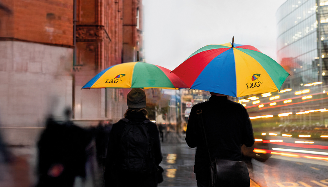 Two people walking through a rainy city street under brightly coloured L&G branded umbrellas, with blurred lights and buildings in the background.