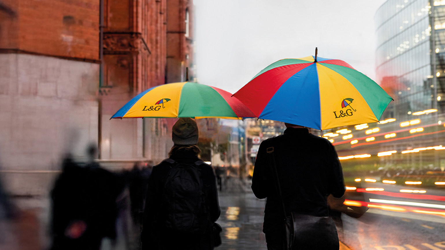 Two people walking through a rainy city street under brightly coloured L&G branded umbrellas, with blurred lights and buildings in the background.
