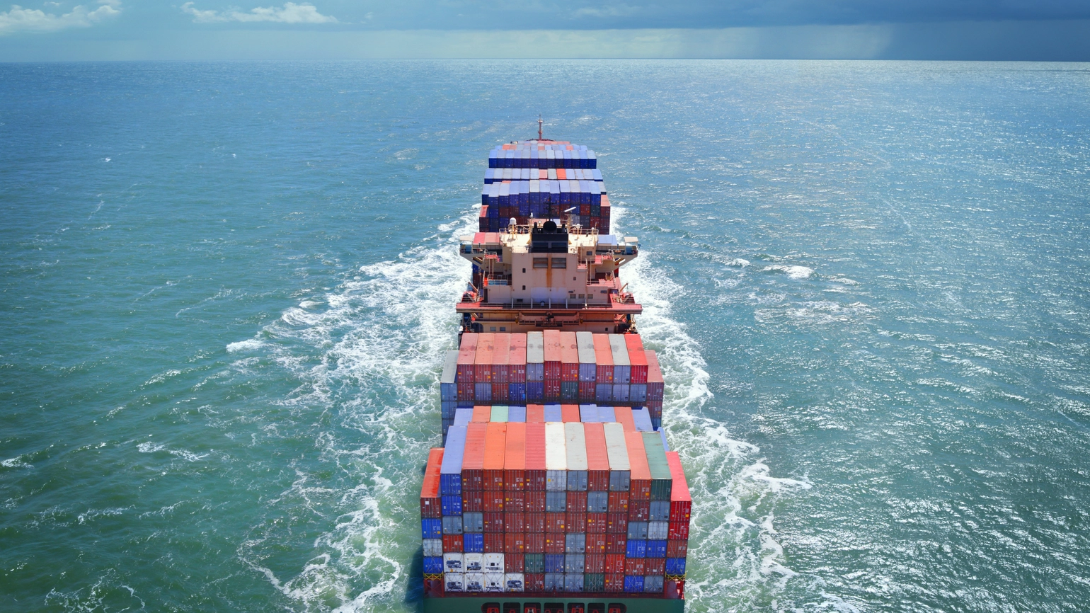 Aerial view of a large freight ship carrying red, blue, and white cargo containers, sailing through the open sea. The ship creates a wave as it moves forward, with water stretching to the horizon under a partly cloudy sky. Aerial view of a large freight ship carrying red, blue, and white cargo containers, sailing through the open sea. The ship creates a wave as it moves forward, with water stretching to the horizon under a partly cloudy sky.