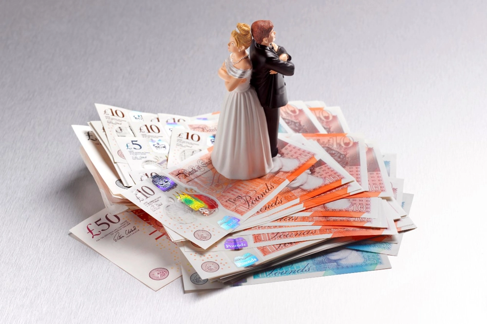 Wedding cake topper of a bride and groom standing back-to-back with arms crossed, placed on a stack of banknotes. Wedding cake topper of a bride and groom standing back-to-back with arms crossed, placed on a stack of banknotes.