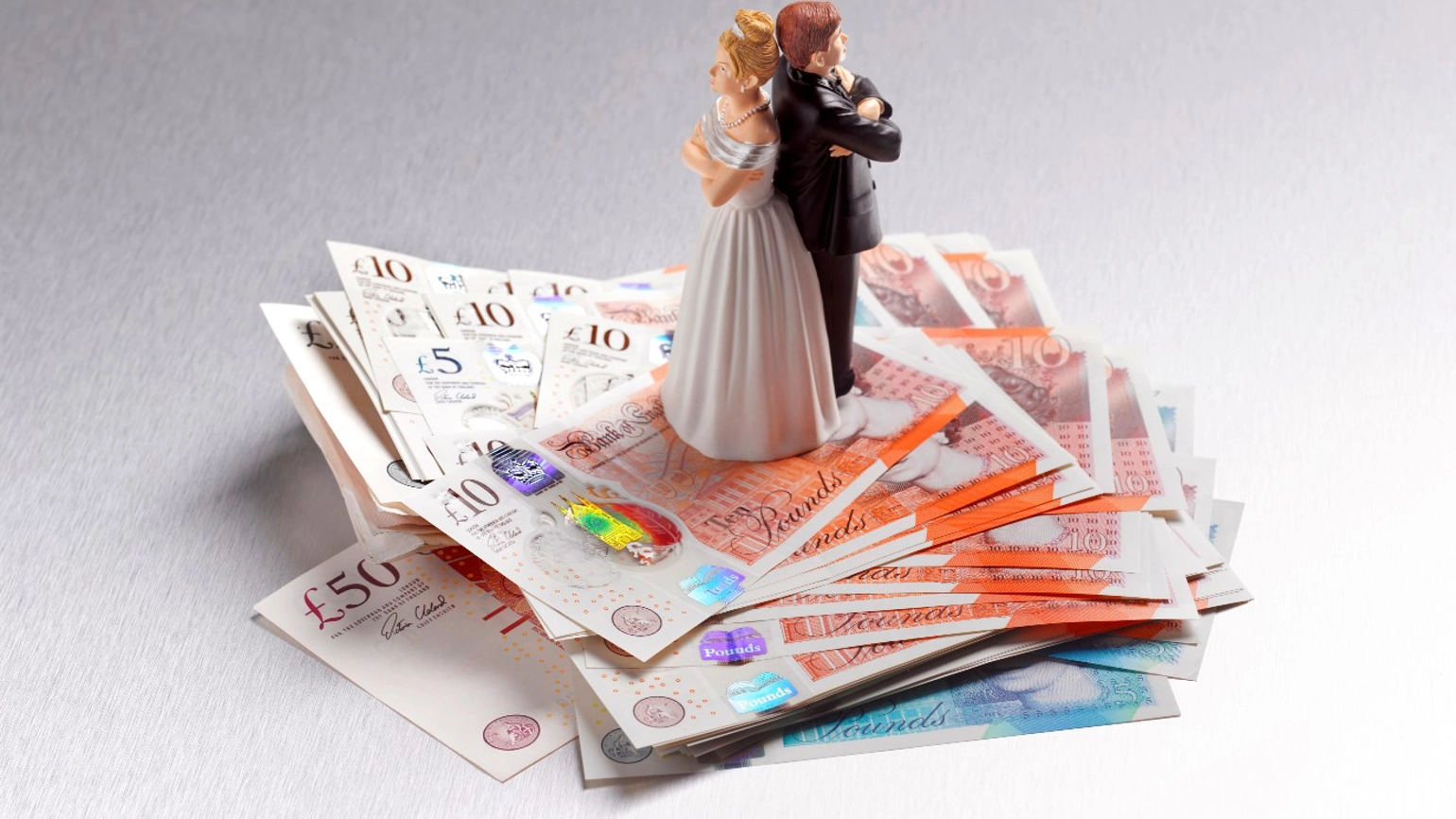 A wedding cake topper of a bride and groom, with backs turned and arms crossed, sits on top of a pile of bank notes A wedding cake topper of a bride and groom, with backs turned and arms crossed, sits on top of a pile of bank notes
