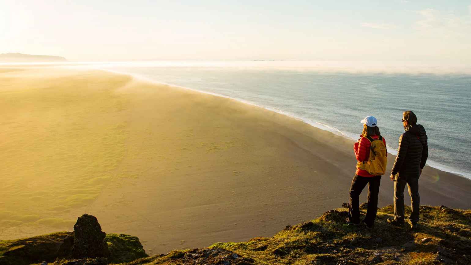 A man and a woman stand on a hill with their backs to the camera, gazing out at a sandy beach that stretches before them. The sun is beginning to set, casting a warm, golden light.