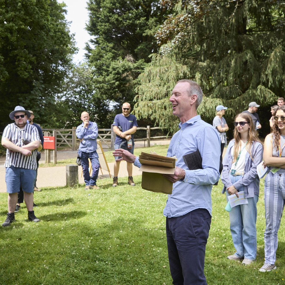 A group of people gathered outdoors on a grassy area with trees in the background, listening to a person holding papers and gesturing while speaking. A group of people gathered outdoors on a grassy area with trees in the background, listening to a person holding papers and gesturing while speaking.