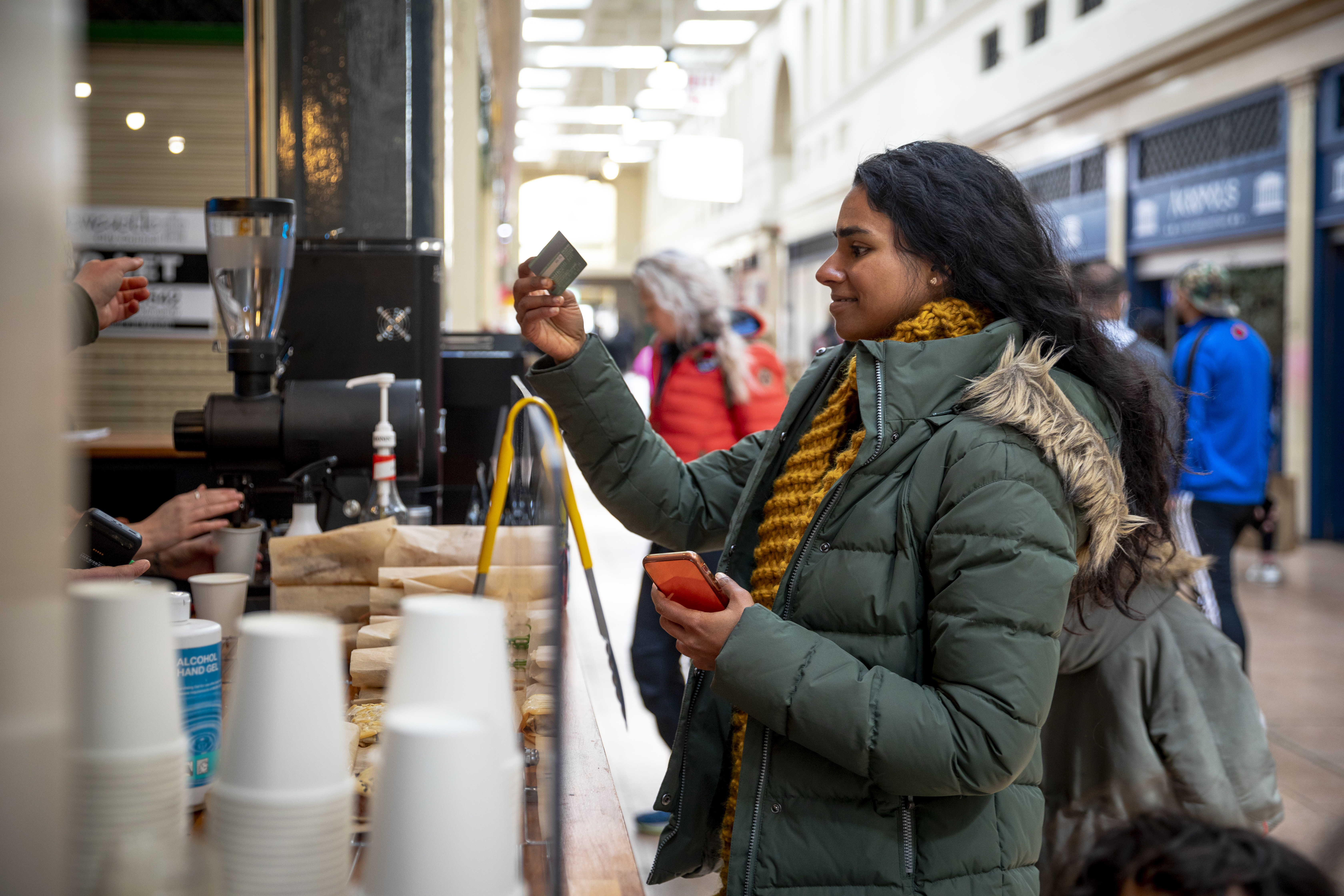 A mid adult woman on a sunny winters day. She is wearing casual, winter clothing and accessories. She is paying for a coffee with her credit card at a small coffee shop in a shopping arcade.