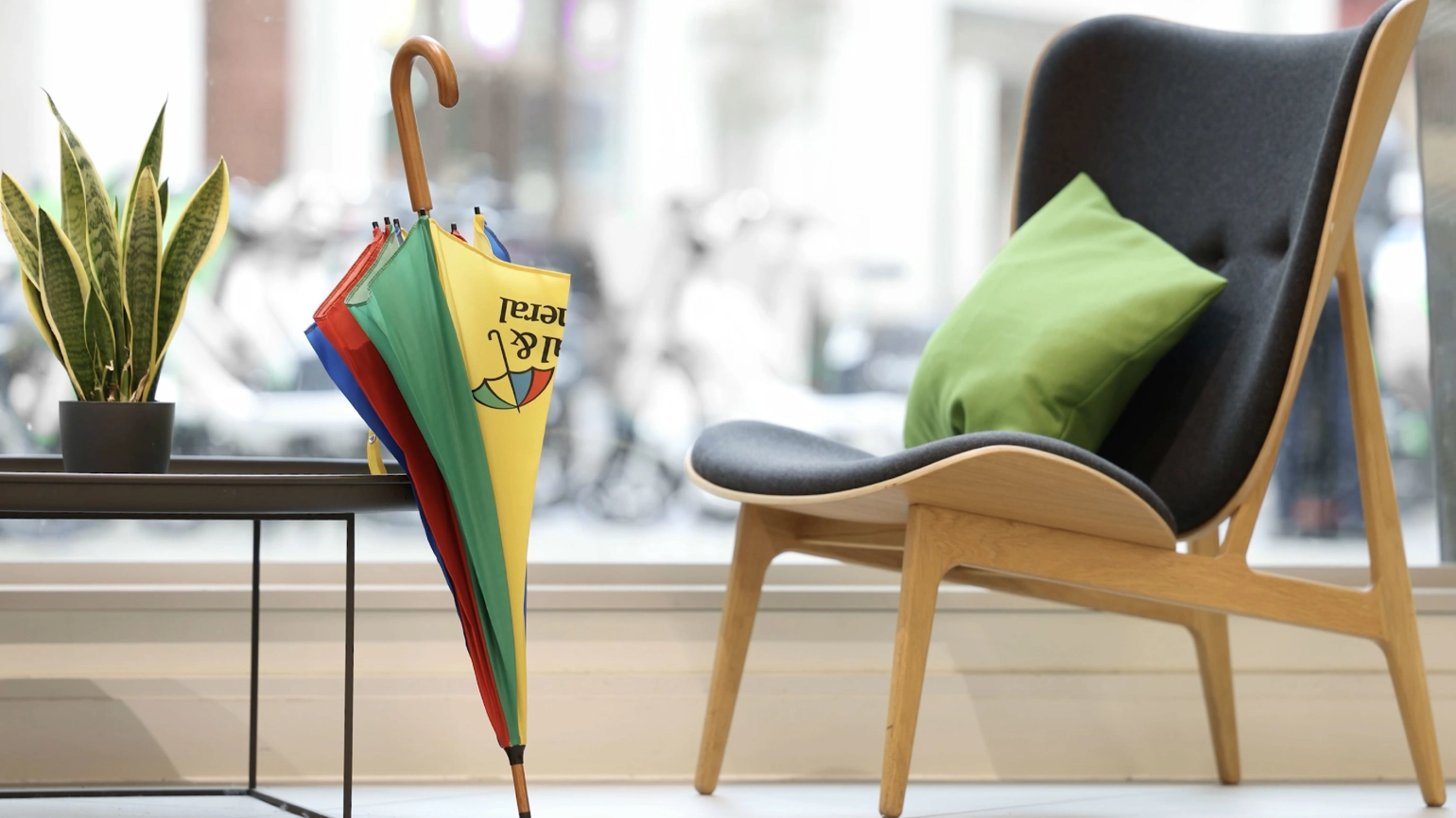 A wooden chair with dark upholstery and a green cushion in front of a large window. Next to the chair is a small black table with a potted plant. On the floor, leaning against the table, is a colourful umbrella with yellow, green, red, and blue panels. The Legal & General logo is visible on the yellow panel. A wooden chair with dark upholstery and a green cushion in front of a large window. Next to the chair is a small black table with a potted plant. On the floor, leaning against the table, is a colourful umbrella with yellow, green, red, and blue panels. The Legal & General logo is visible on the yellow panel.