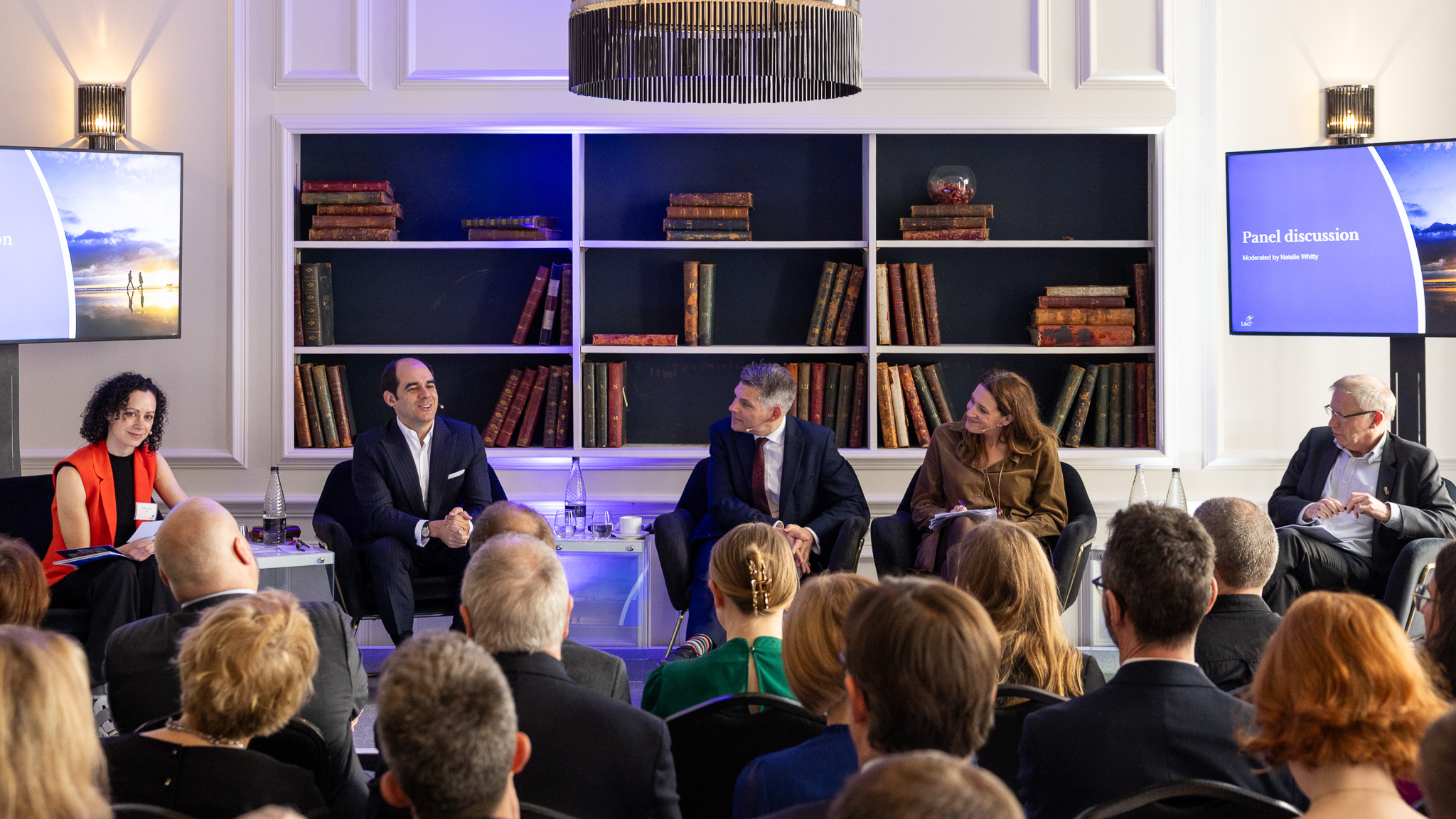 Panel of five speakers seated on stage in front of bookshelves while an audience watches the discussion.