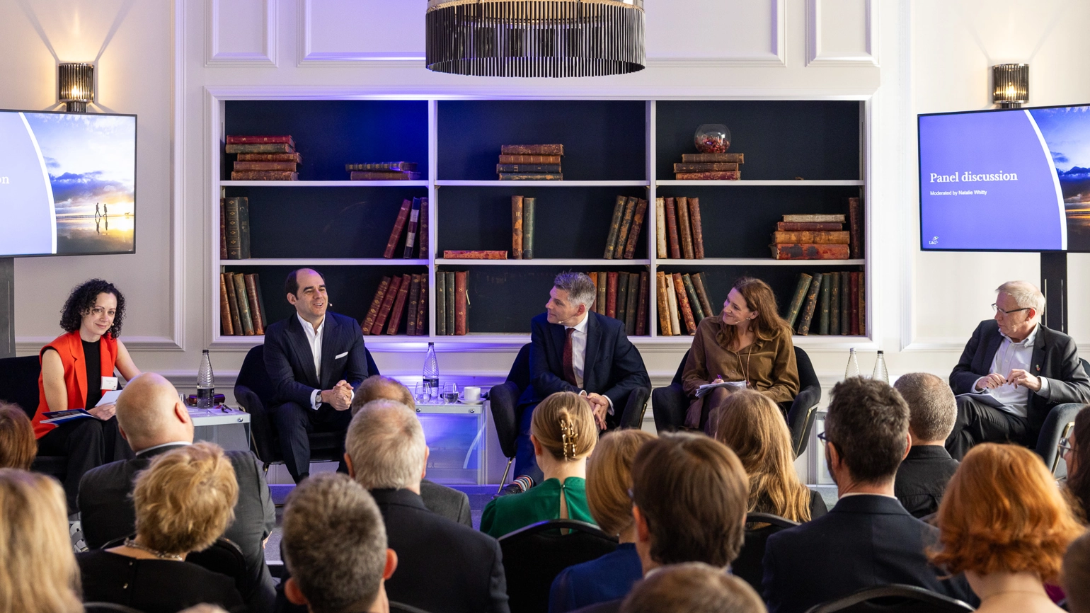 Panel of five speakers seated on stage in front of bookshelves while an audience watches the discussion. Panel of five speakers seated on stage in front of bookshelves while an audience watches the discussion.